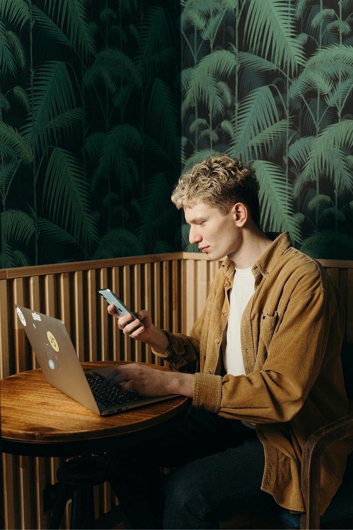 Young man with curly blond hair sitting at a wooden café table, multitasking between a laptop and a smartphone, with a leafy tropical wallpaper in the background.